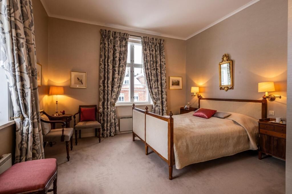 A bedroom with a four-poster bed, two bedside tables with lamps, a chair, a small table, and a window with curtains.