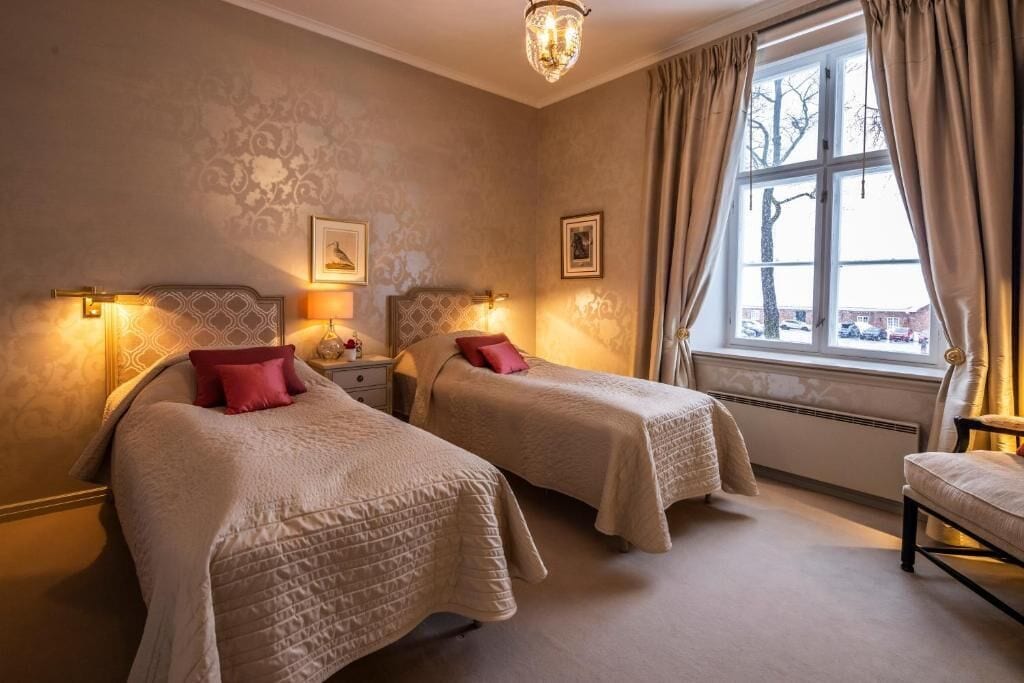 Una habitación de hotel con dos camas, un candelabro, una ventana con cortinas e imágenes enmarcadas en la pared.
