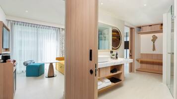 A modern hotel room with a wooden wardrobe, a bathroom with a large mirror, a sink, and a shower area.