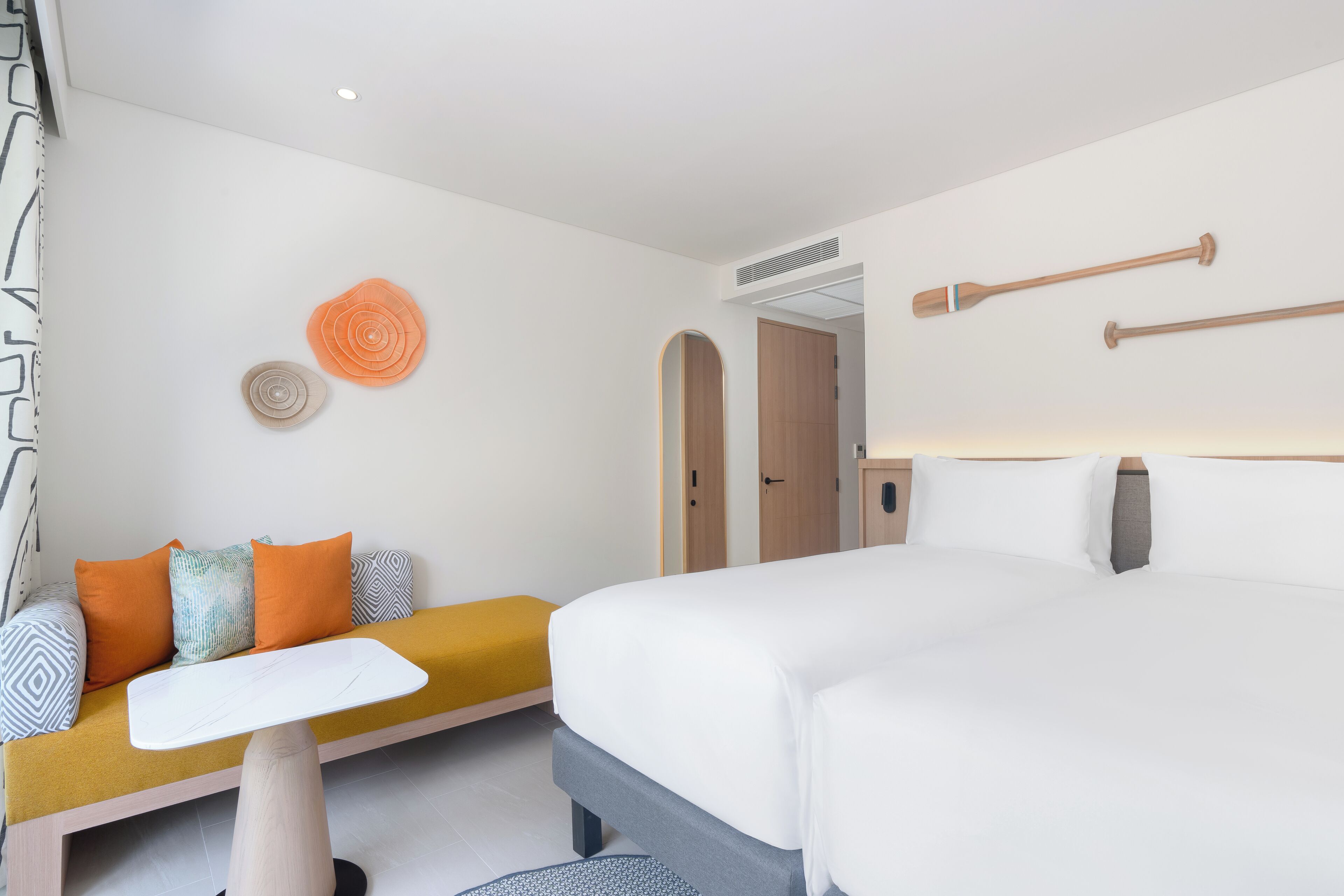 A modern hotel room with a large bed, a sofa with orange cushions, a small table, and decorative wall art.