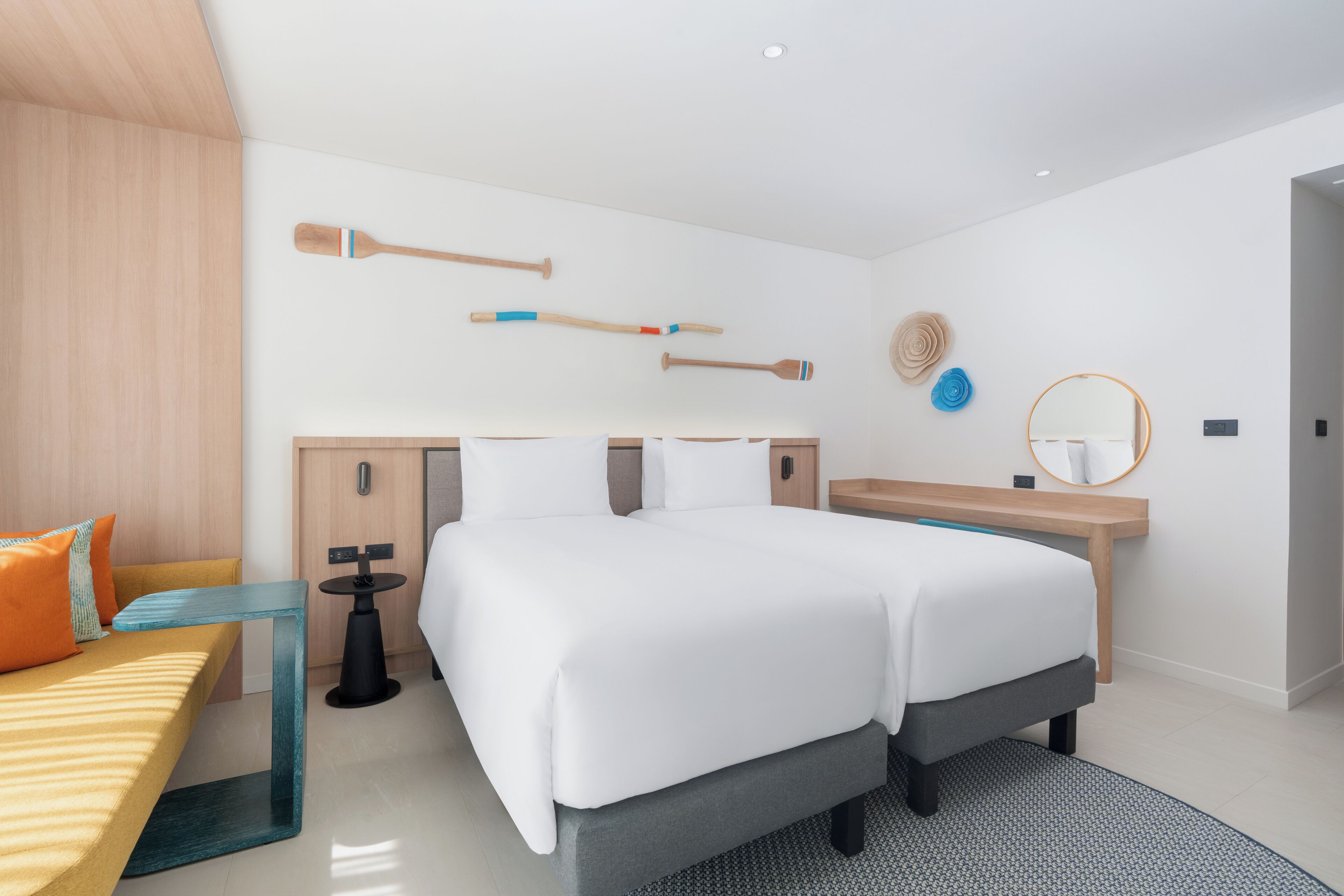 A modern hotel room with a large bed, a wooden headboard, a bedside table, a bench, and a wall-mounted mirror.