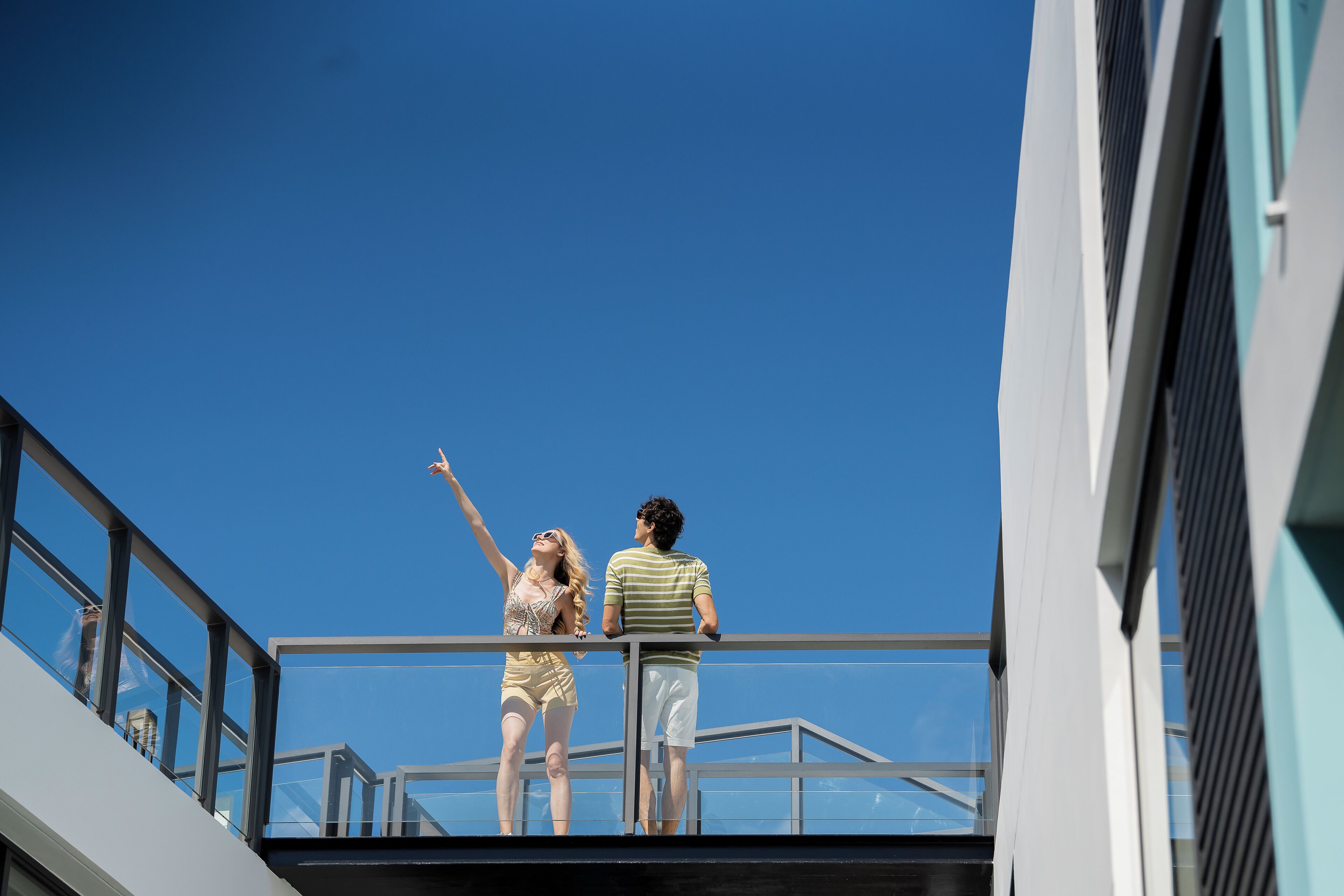 Dos personas de pie en un balcón con un cielo azul despejado al fondo.