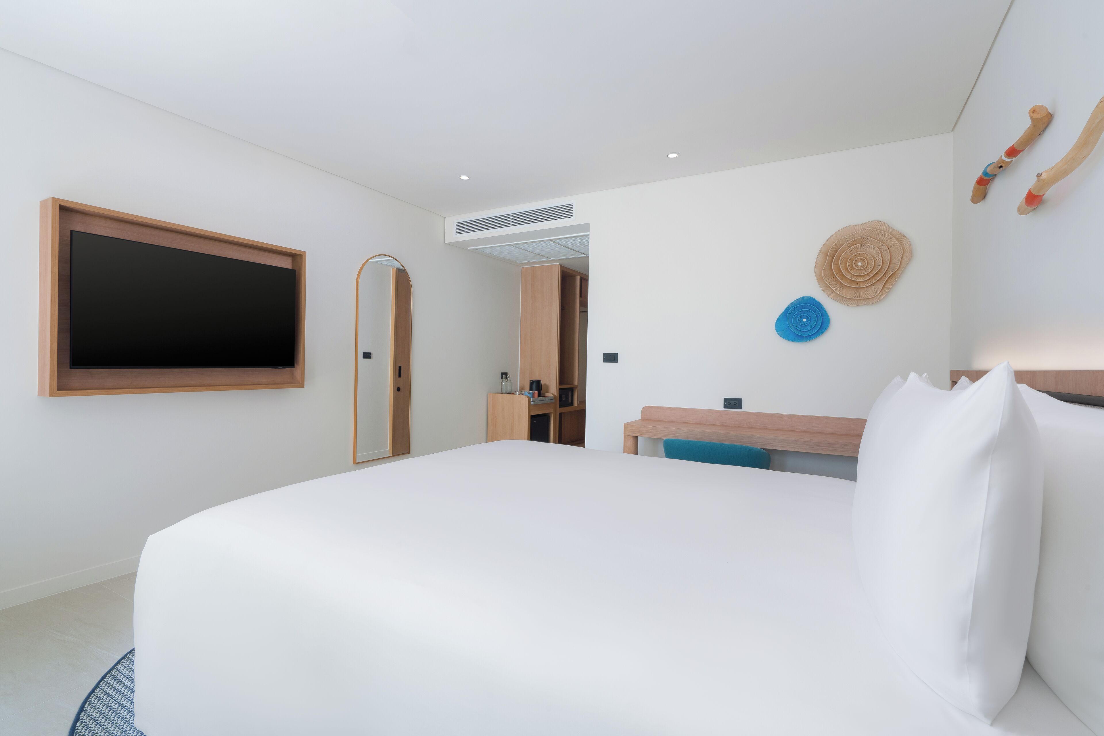 A modern hotel room with a large bed, a flat-screen TV mounted on the wall, a wooden desk, and a small seating area.