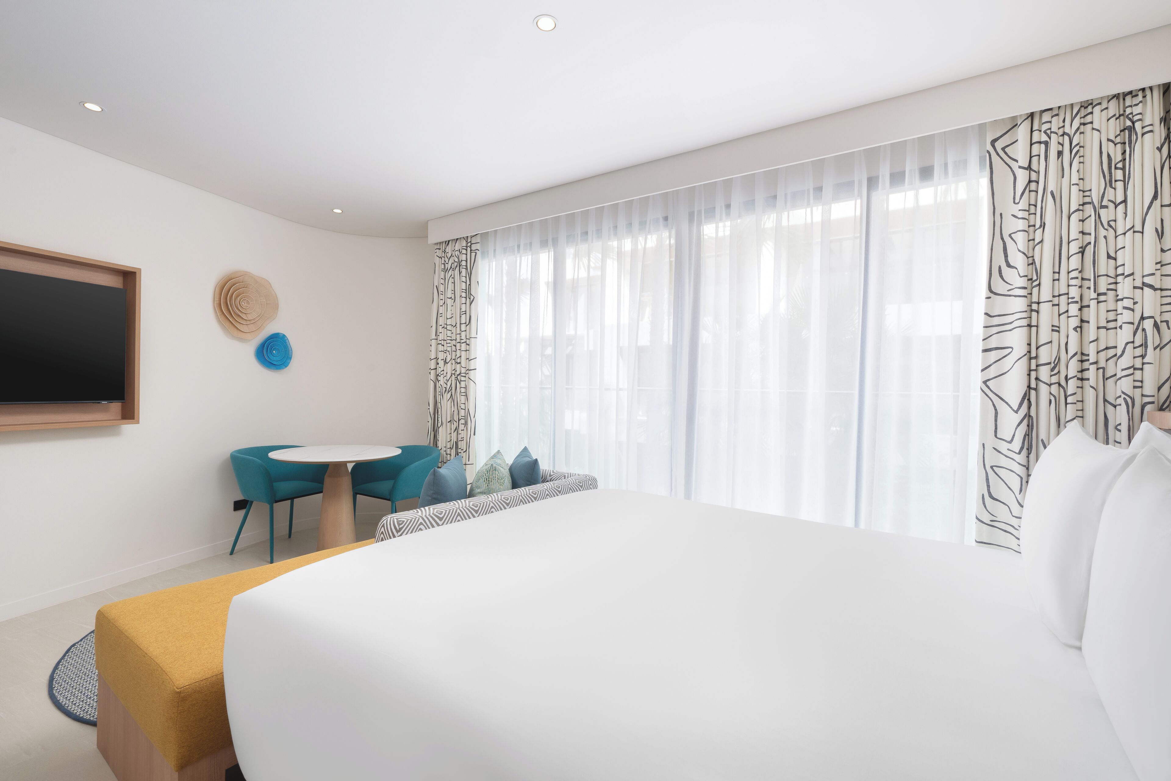 A modern hotel room with a large bed, a round table with chairs, and a view of the outdoors through sheer curtains.