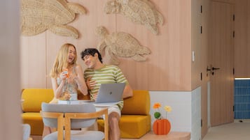 A modern living room with a yellow sofa, a wooden coffee table, and a laptop on the sofa. There are decorative wall art pieces and a vase with flowers on the coffee table.