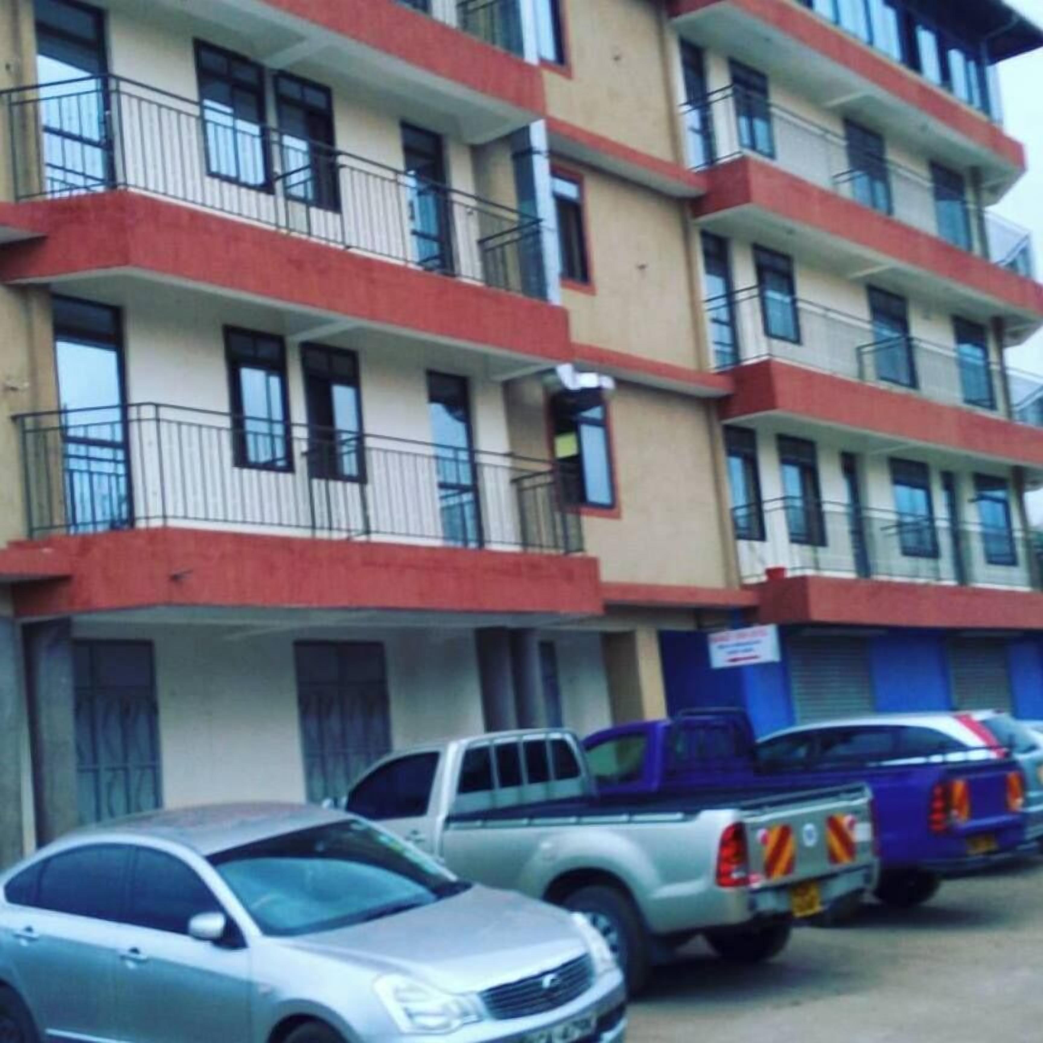 A multi-story building with balconies, parked vehicles, and a sign indicating a commercial space.