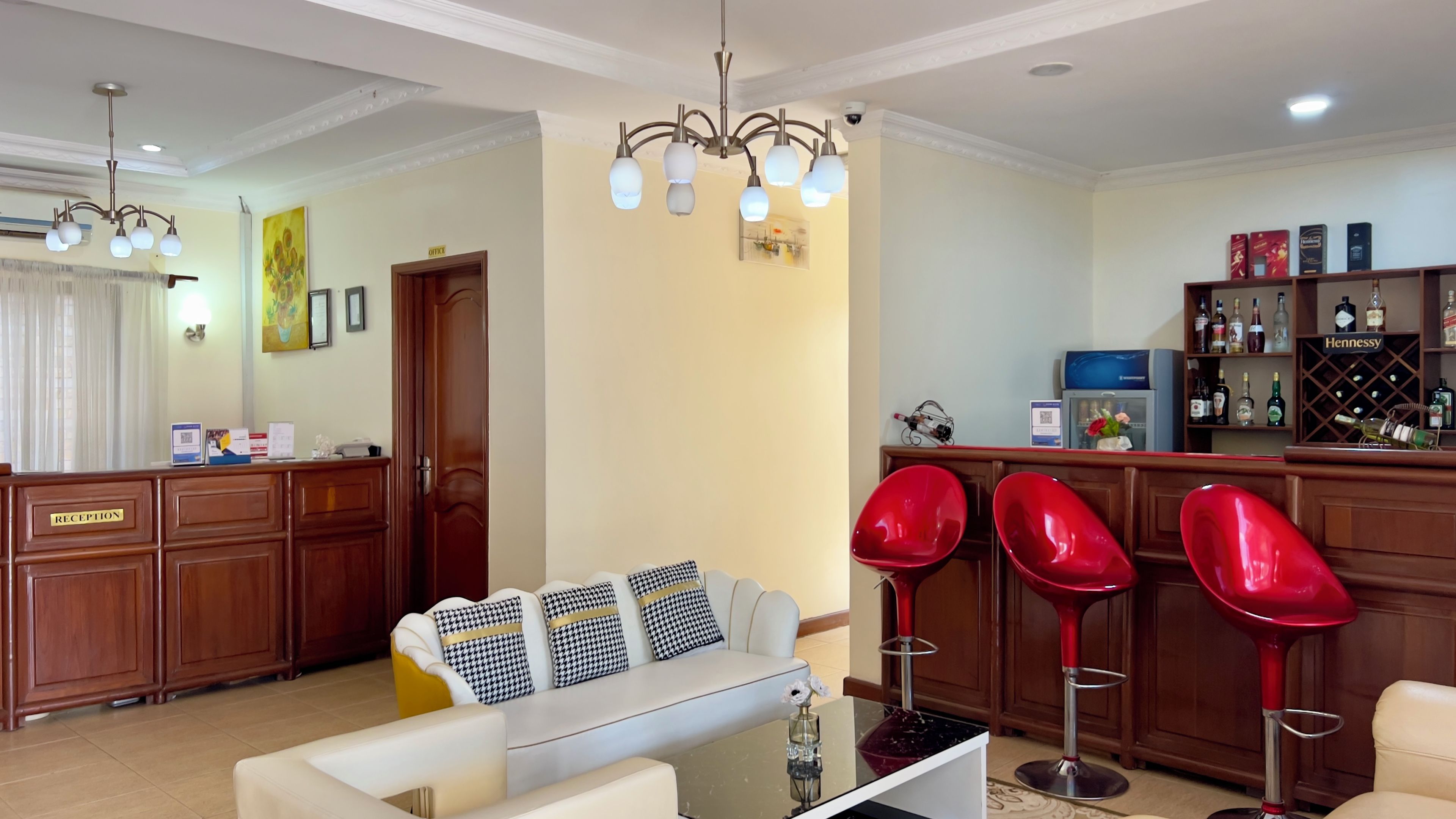 A hotel reception area with a reception desk, a sofa with patterned cushions, a bar area with red bar stools, and a wine display.
