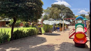 A playground with slides and swings, surrounded by trees and a brick building. - Explora Lodge (Arusha)