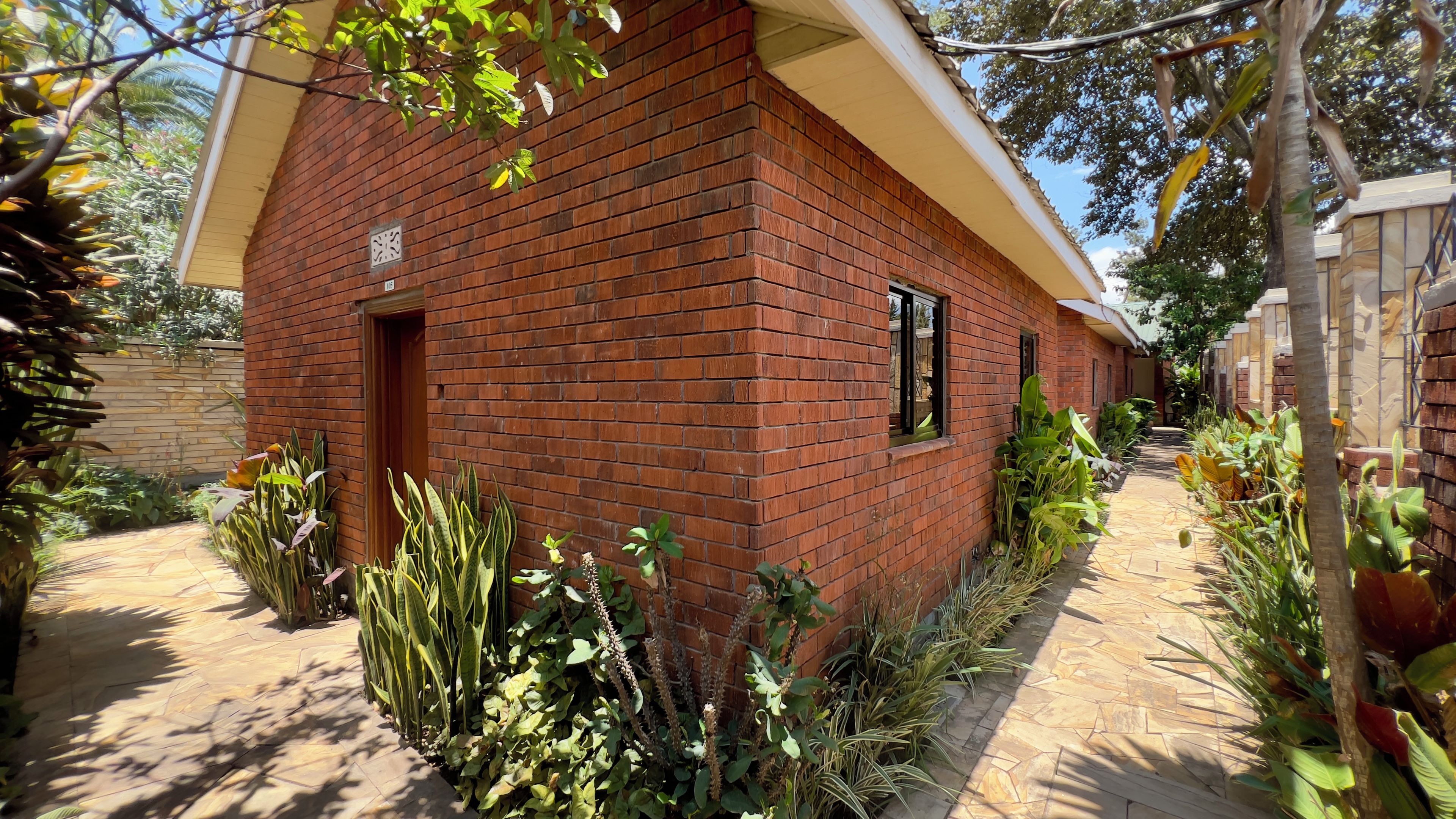 A brick building with a door and windows, surrounded by various plants and a paved walkway.