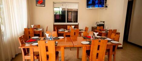 A dining area with wooden tables and chairs, a large window, and a television mounted on the wall.