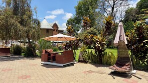 An outdoor seating area with a table and chairs, surrounded by lush greenery and a building in the background. - Explora Lodge (Arusha)