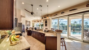 Une cuisine spacieuse avec un grand îlot central, un coin repas offrant une vue sur la piscine et un ventilateur de plafond.