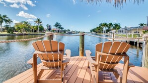 Une terrasse en bois avec deux fauteuils Adirondack donnant sur une marina où sont amarrés des bateaux, avec des palmiers en arrière-plan.