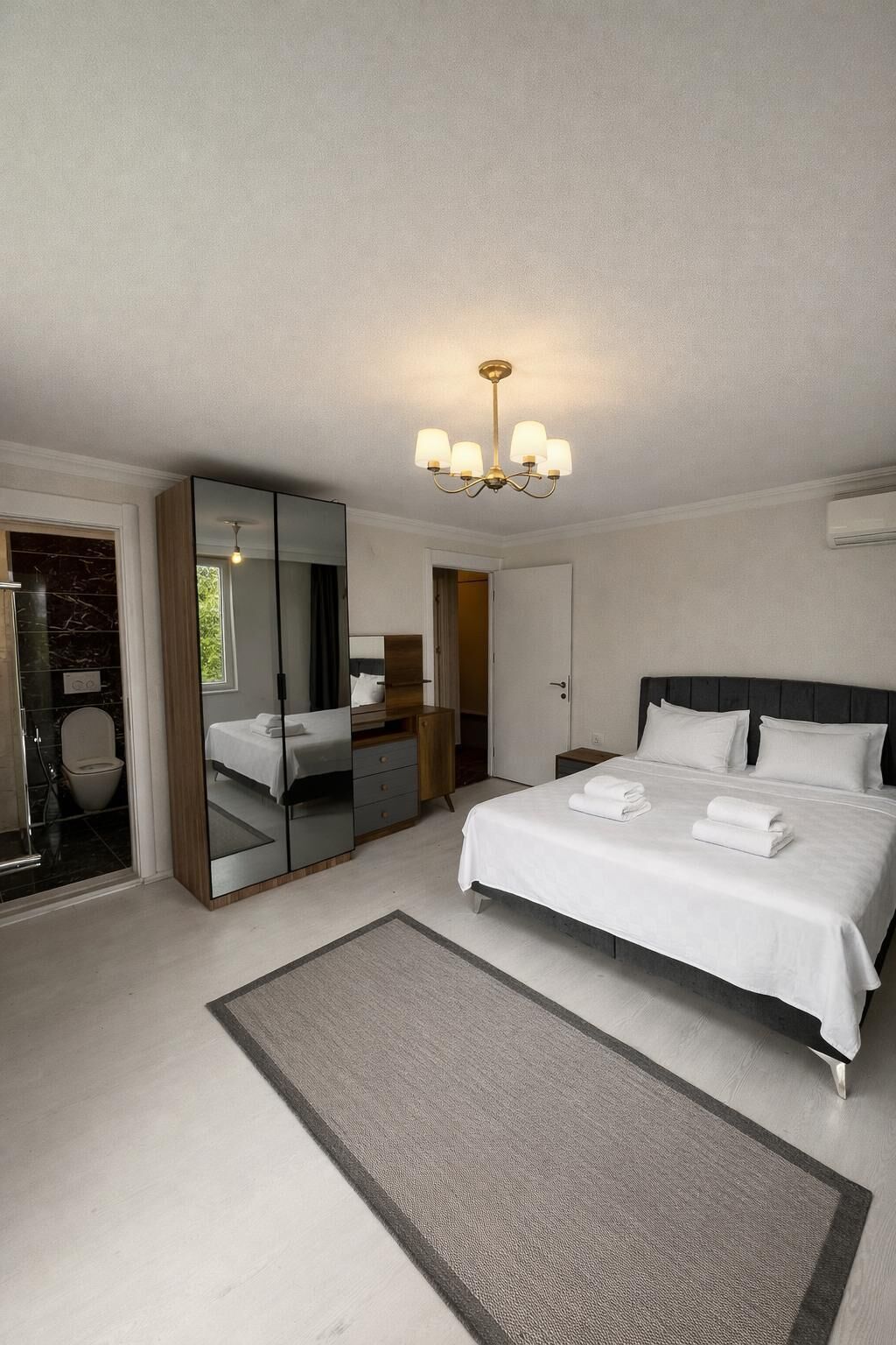 A modern hotel room with a large bed, a mirror, a chandelier, and a bathroom with a marble shower.