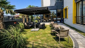 An outdoor patio with seating, a covered area, and a building with large windows and yellow accents. - Luxury 3BR Apartment with Mountain View, AC & Pool – Maşukiye (Kocaeli)