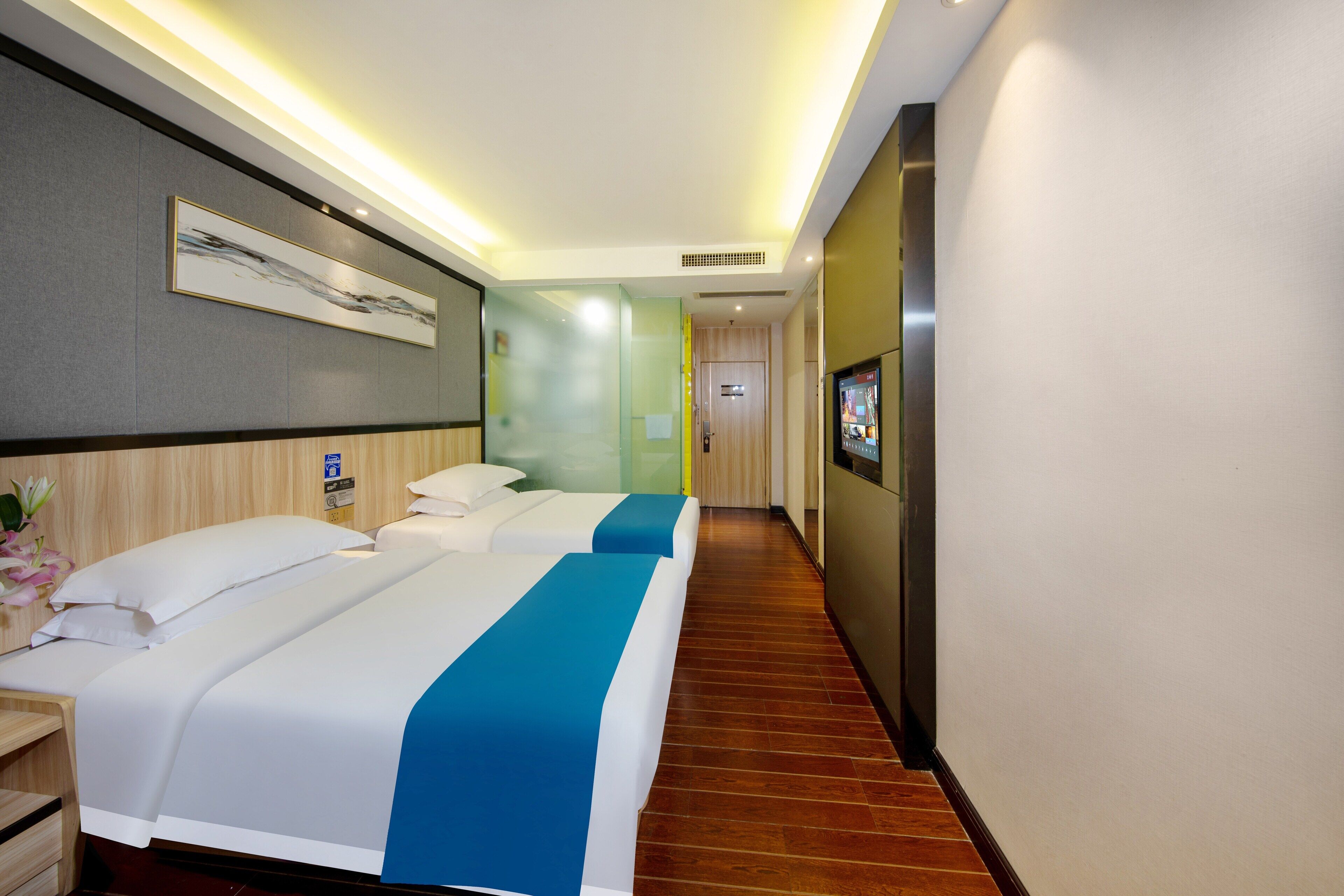 Habitación de hotel con dos camas, suelo de madera, televisor montado en la pared y una obra de arte enmarcada.