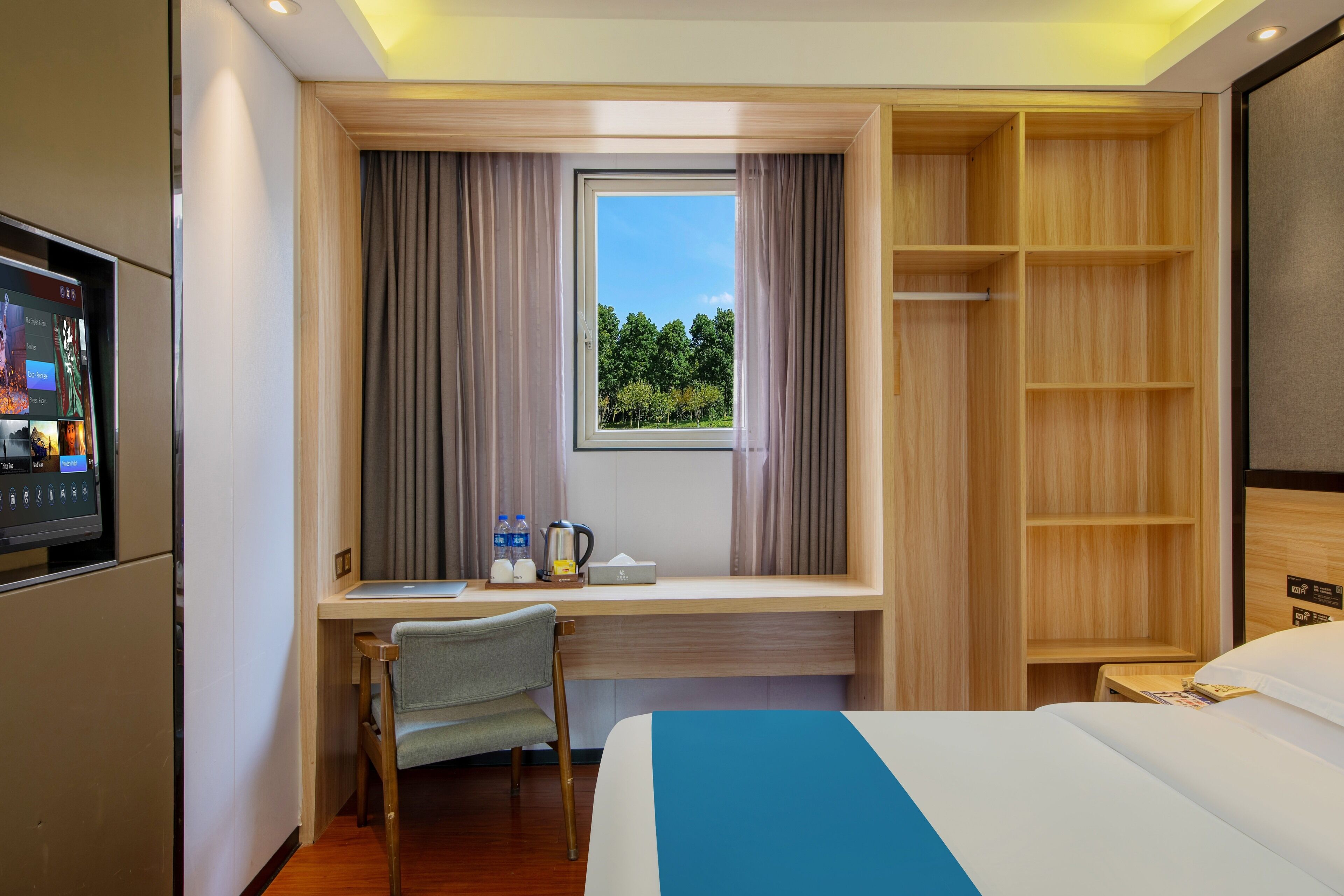 A hotel room with a bed, a desk with a chair, a window with curtains, a TV, and a wooden wardrobe.