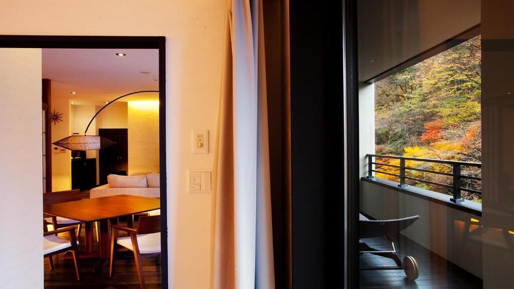 Un interior moderno con comedor, sofá y vistas al follaje otoñal desde un balcón.