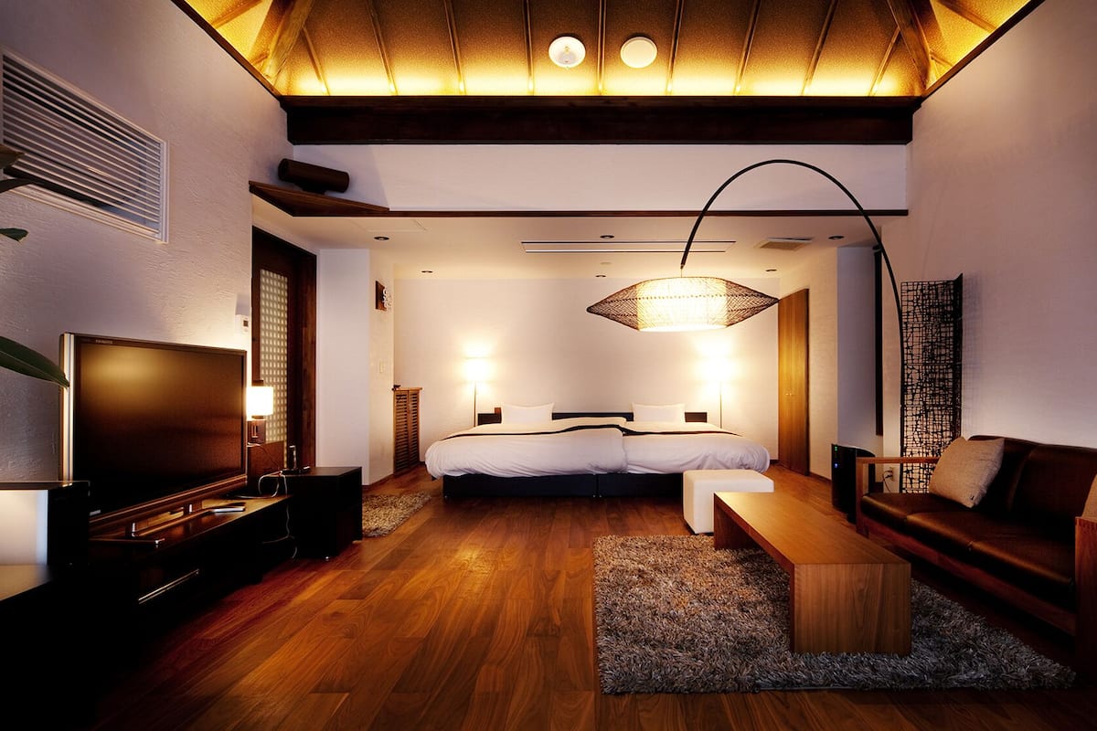 A modern hotel room with a wooden floor, a large bed, a sofa, a TV, and a ceiling light fixture.