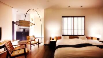 A hotel room with a bed, a desk with a chair, a hanging light fixture, and a window with vertical blinds.