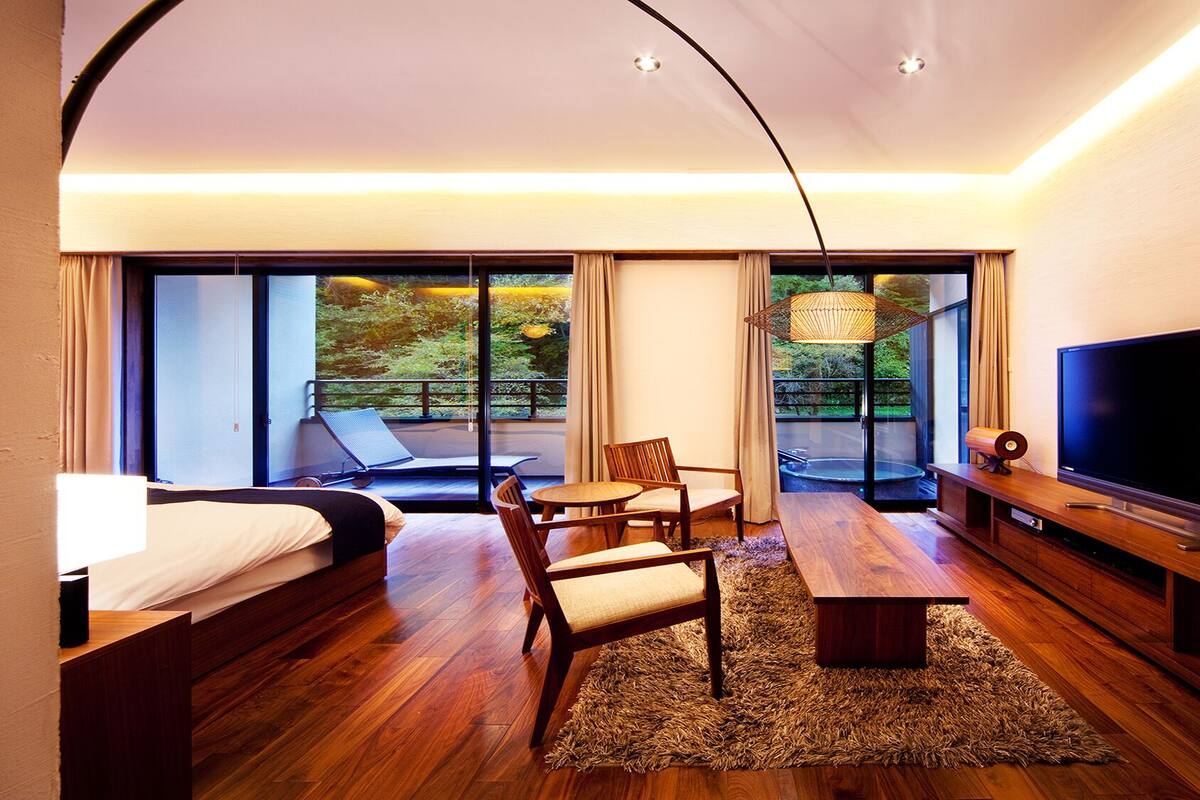 A modern hotel room with a bed, a dining table, chairs, a TV, and large windows offering a view of greenery.