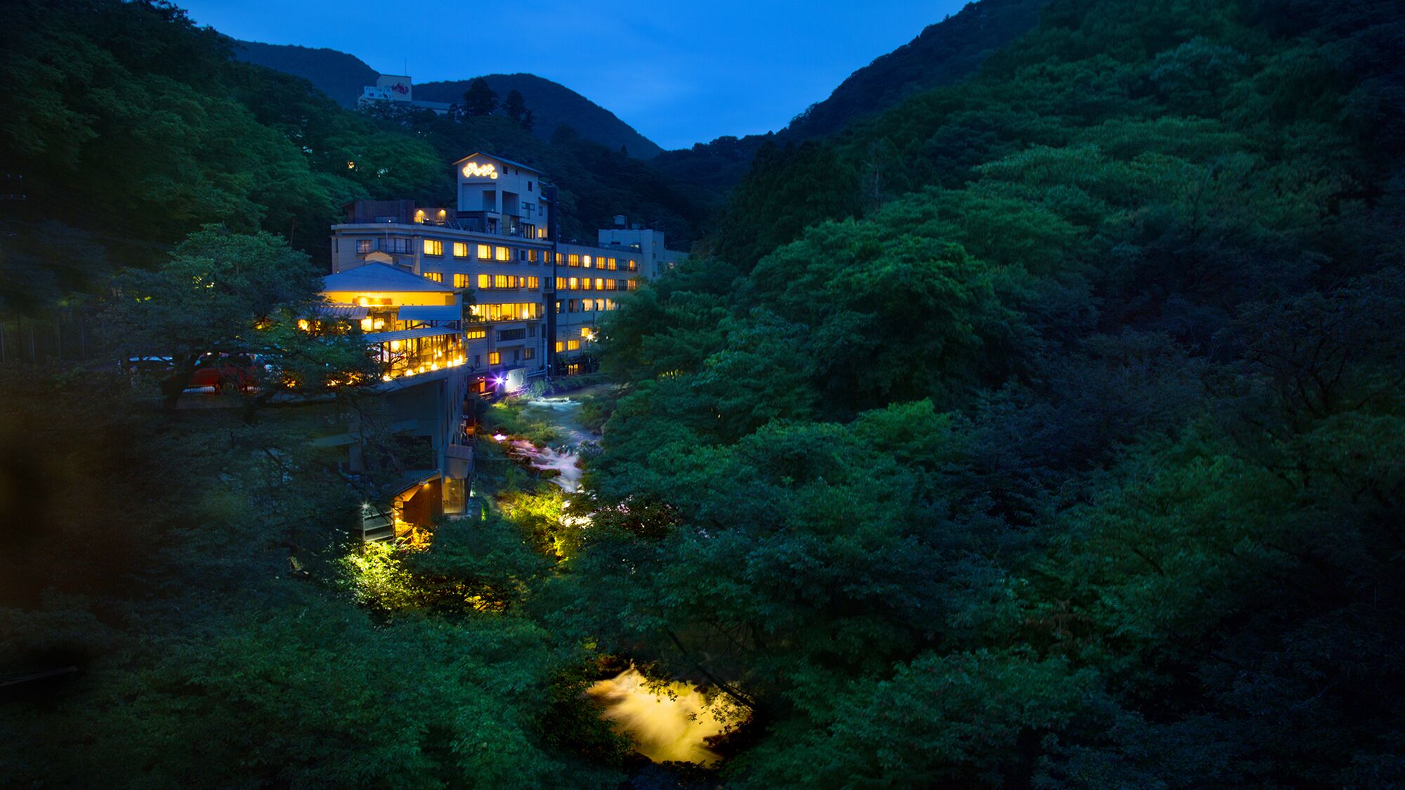 Un hotel de varios pisos con un río que atraviesa la propiedad, rodeado de un bosque denso y colinas.
