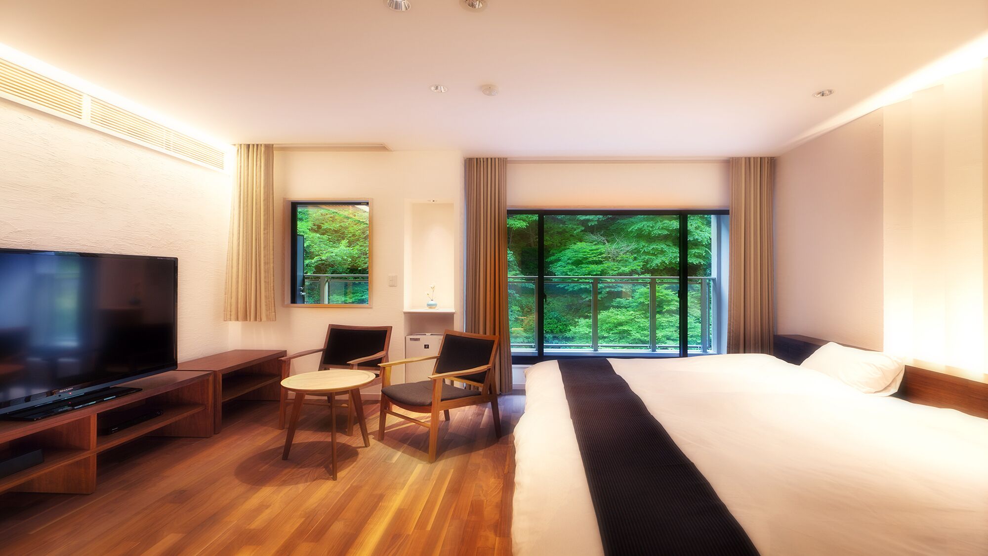 Une chambre d’hôtel avec un grand lit, une télévision, un bureau et une chaise. Il y a une fenêtre donnant sur des arbres.
