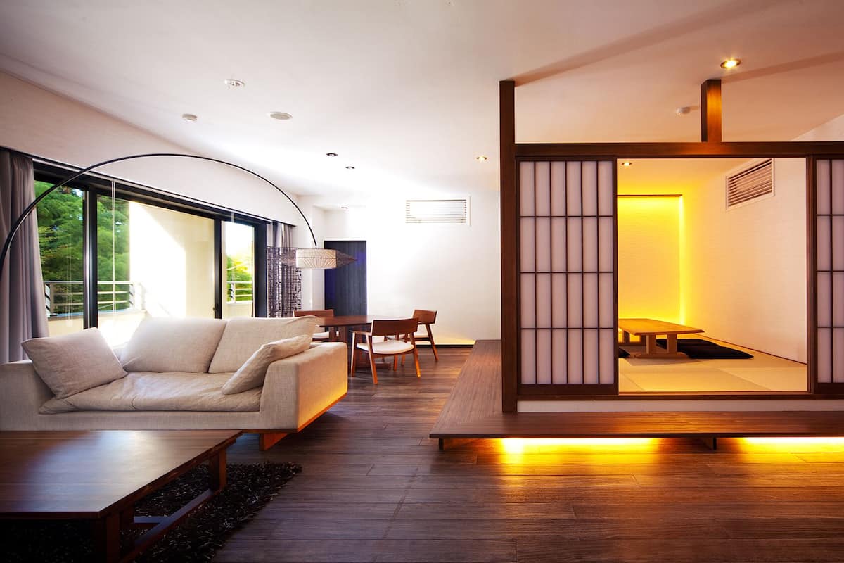 A modern living room with a sofa, dining table, and chairs. There is a large window and a view of greenery outside.
