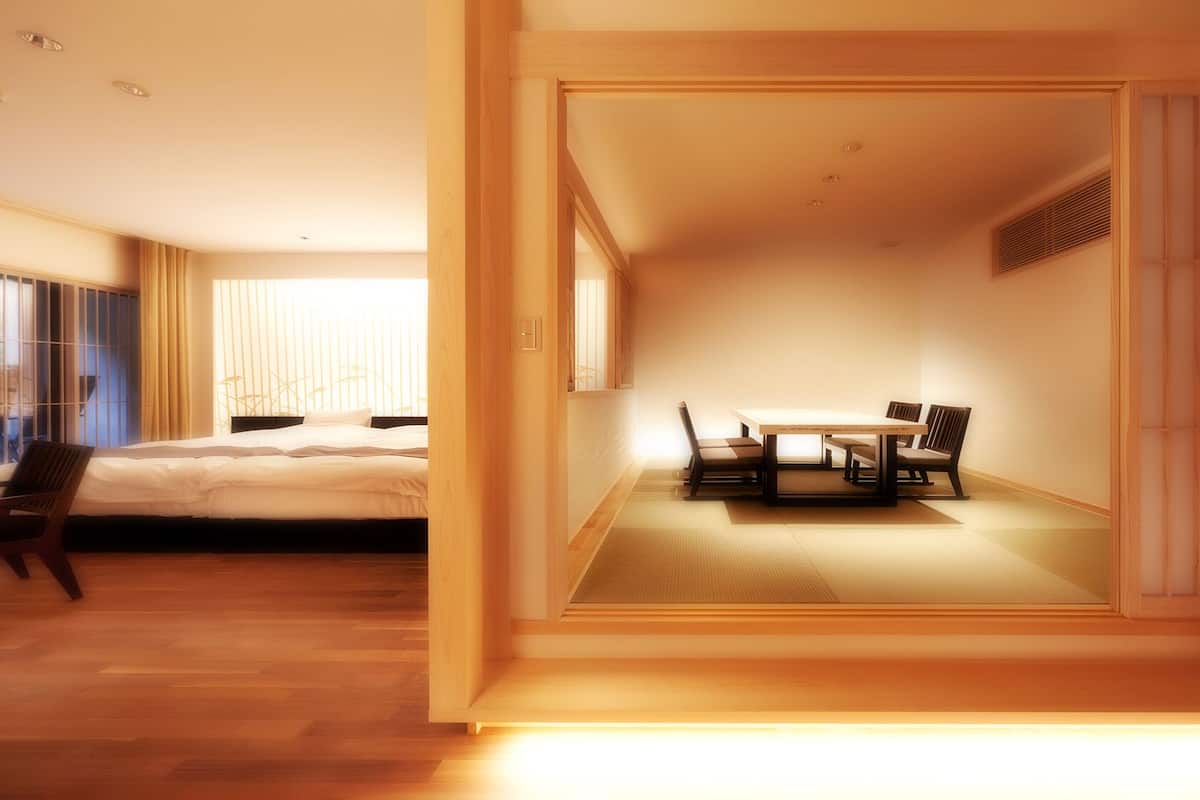 A modern hotel room with a bed, a dining area with a table and chairs, and a sliding door leading to another room.