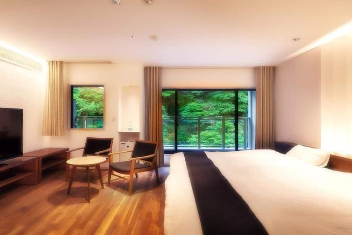 A hotel room with a large bed, a TV, a desk with a chair, and a window with a view of greenery.