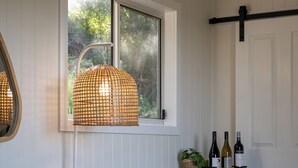 A room with a white chair, a small table with bottles, a wicker lamp, and a window with a view of trees.