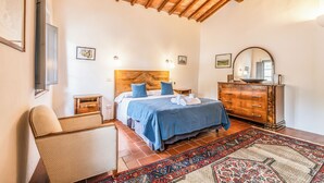 A bedroom with a wooden headboard, a bed with blue bedding, a dresser, a chair, and a patterned rug. - Tenuta della Selva - Castellaccio 7 (Monteroni d'Arbia)
