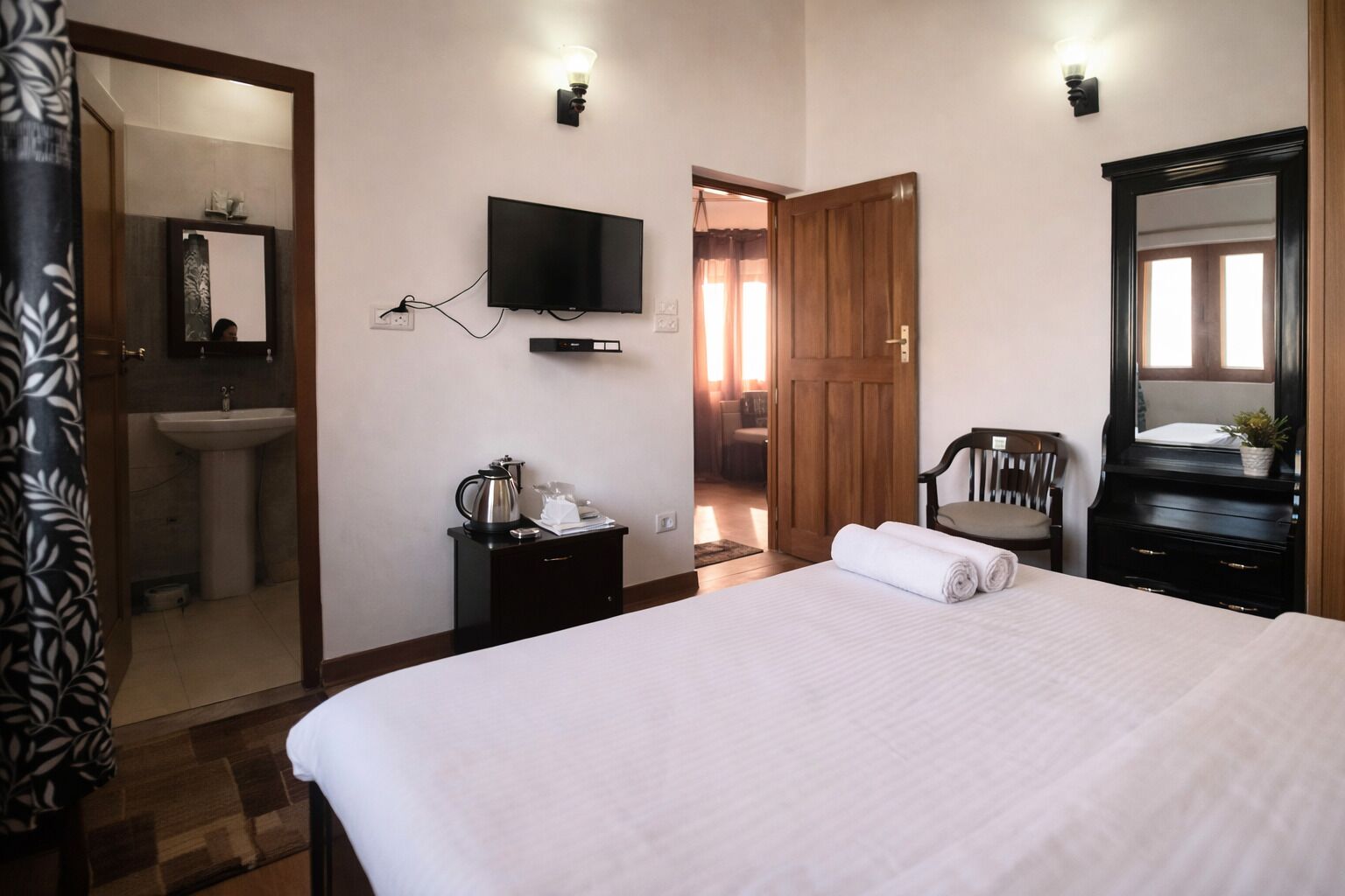A hotel room with a bed, a TV mounted on the wall, a wooden door, a mirror, a small table with a kettle, and a bathroom visible through an open door.