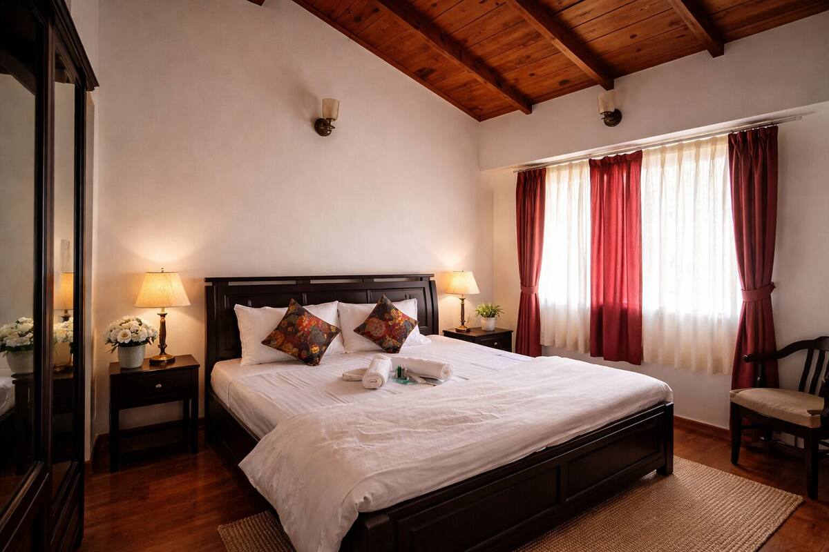 A bedroom with a wooden ceiling, a bed with white bedding, two nightstands with lamps, a chair, and a window with red curtains.