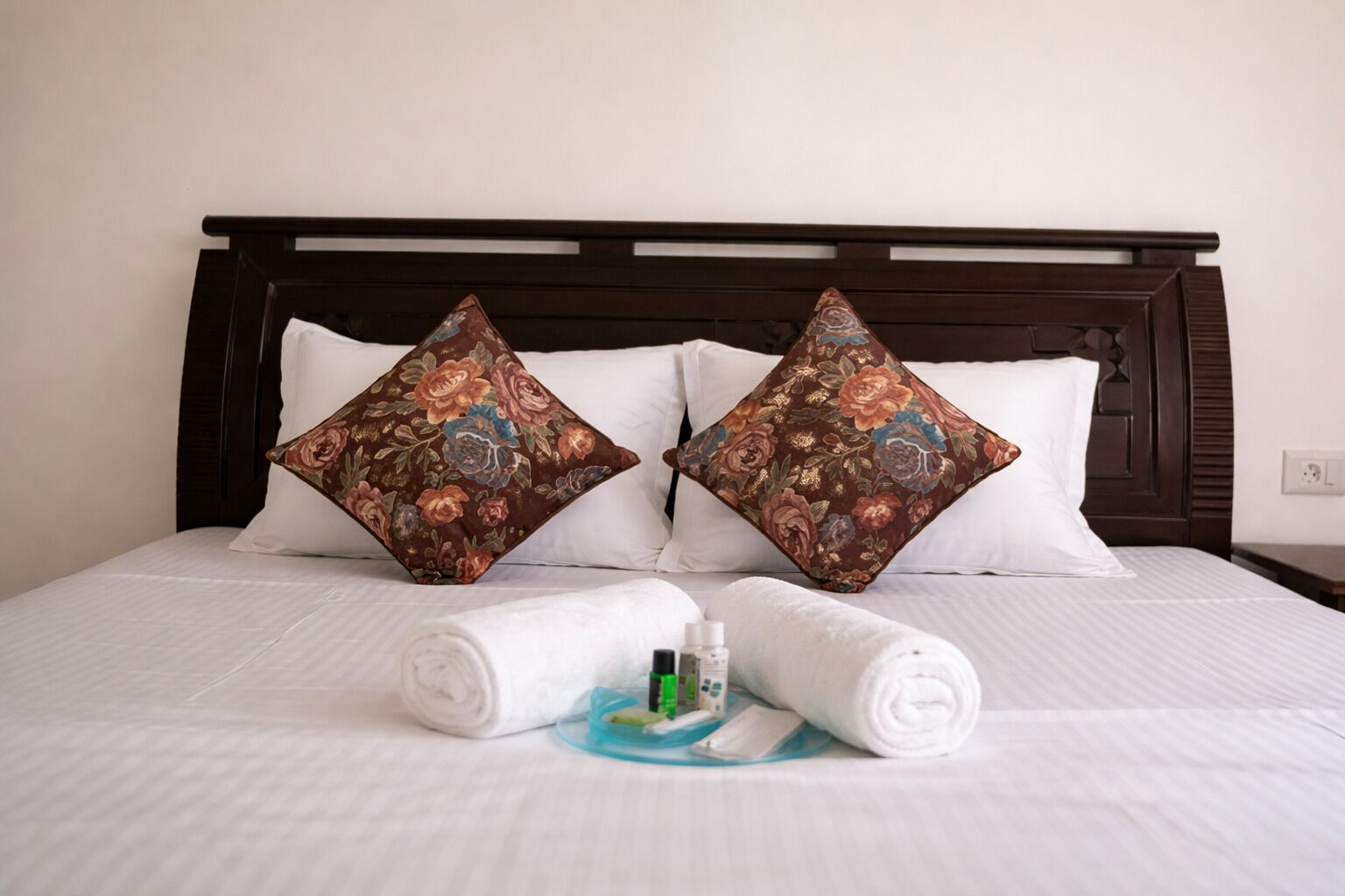 A neatly made bed with floral-patterned pillows, a rolled white towel, and a small bottle of lotion on a blue tray.