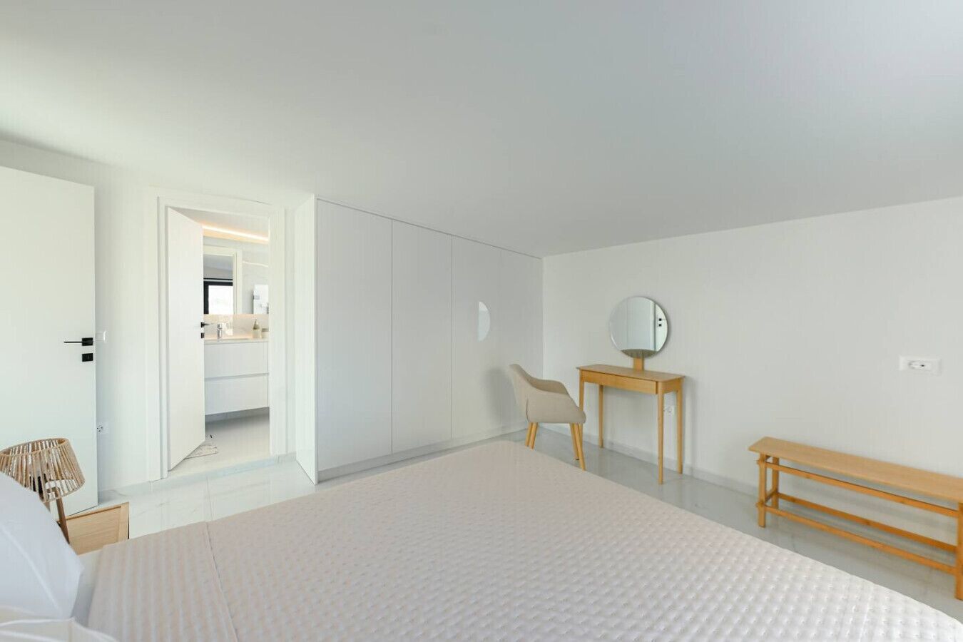 A bedroom with a bed, a wooden bench, a chair, a desk with a mirror, and a bathroom visible through an open door.