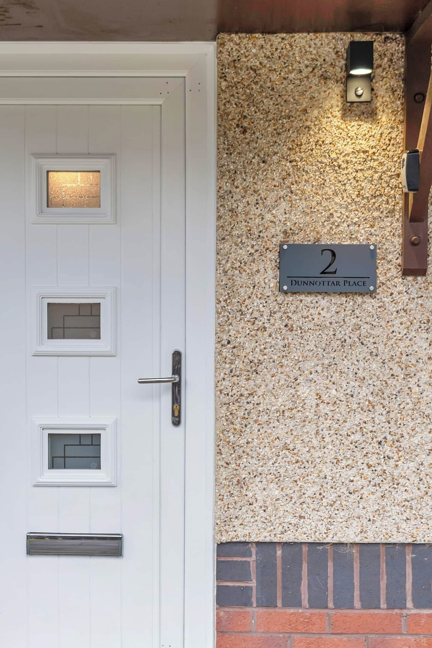 A white door with multiple windows and a metal handle. There is a number plate labeled "2 Dunnottar Place" on a textured wall.
