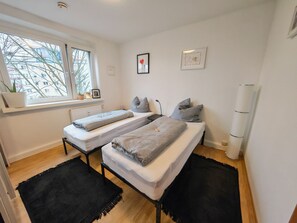 A modern bedroom with two beds, a window with a view of buildings, a radiator, and a bedside table with a lamp.