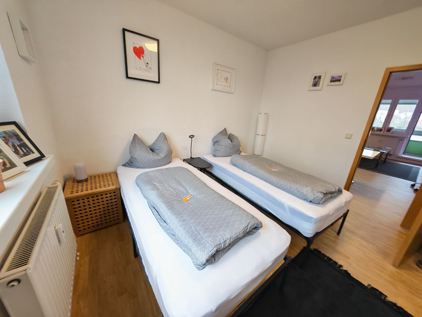 A bedroom with two beds, a radiator, a bedside table, and a door leading to another room.