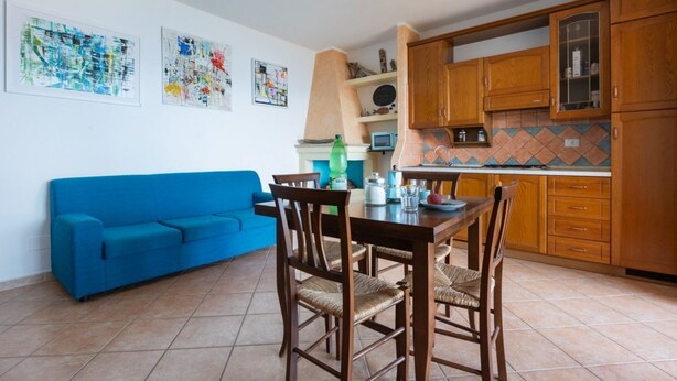 Un salon avec un canapé bleu, une table à manger et des chaises en bois, une cuisine avec des meubles en bois et un dosseret carrelé, et des œuvres d’art abstraites accrochées aux murs.