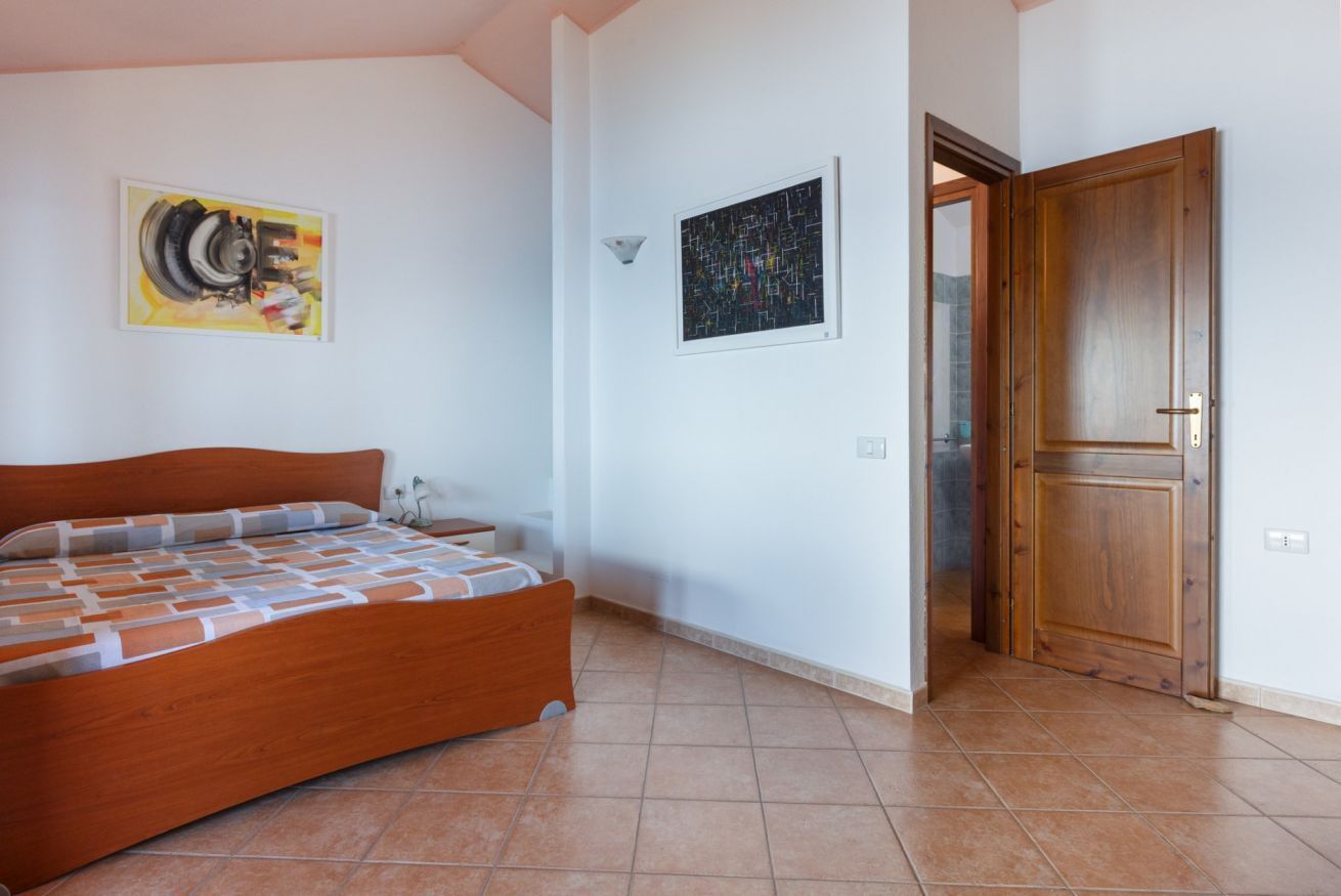 Quarto com cama de madeira, mesa de cabeceira, luminária na parede e porta que dá para outro espaço.