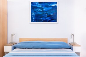 Een slaapkamer met een bed, twee nachtkastjes, een wandlamp, een schilderij met abstracte vormgeving en een houten hoofdbord.
