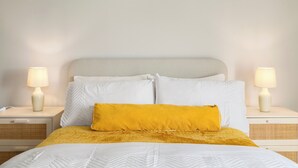 Ein ordentlich bezogenes Bett mit weißer und gelber Bettwäsche, zwei Nachttischlampen und ein schlichter Kopfteil.