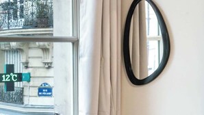Een kamer met een groot raam, witte gordijnen en een spiegel met een zwarte lijst aan de muur.