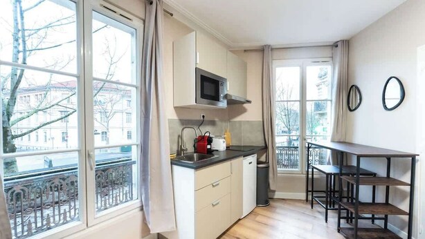 Een compacte keuken met een magnetron, spoelbak en kastjes, grote ramen met uitzicht op gebouwen, en een klein eethoekje met een tafel en stoelen.