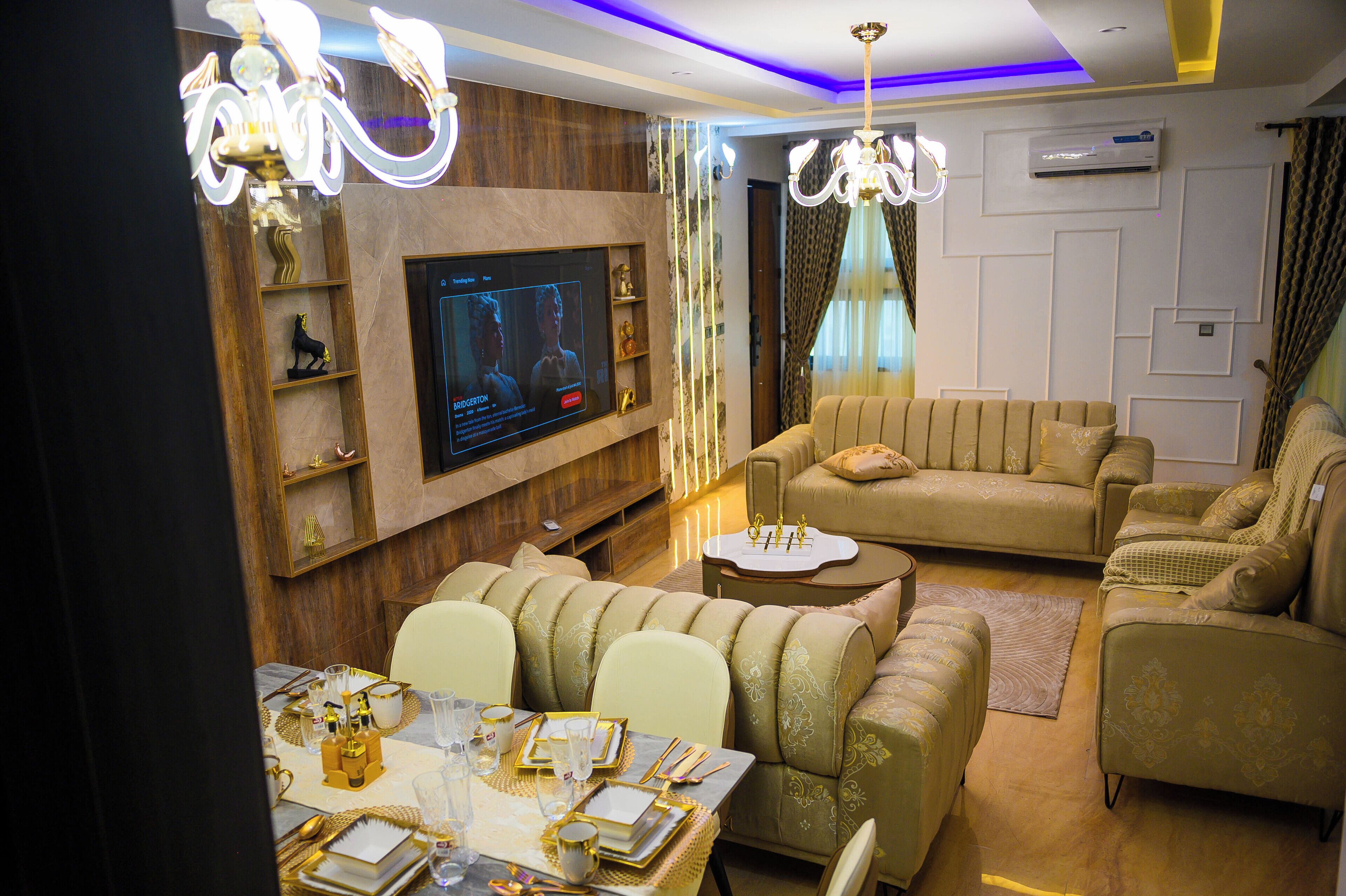 A modern living room with a flat-screen TV, plush sofas, a dining table set for four, and decorative lighting.