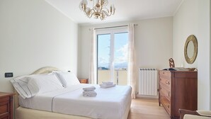 Une chambre à coucher avec un grand lit, une commode en bois, une chaise, un lustre et une vue par la fenêtre.