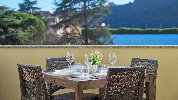 Une table à manger, avec ses chaises, agrémentée d’une plante en pot et de verres à vin, donnant sur un plan d’eau et des collines.