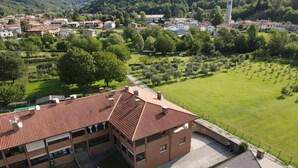 A multi-story building with a red-tiled roof, a parking area with several cars, and a surrounding green landscape with trees and a field.