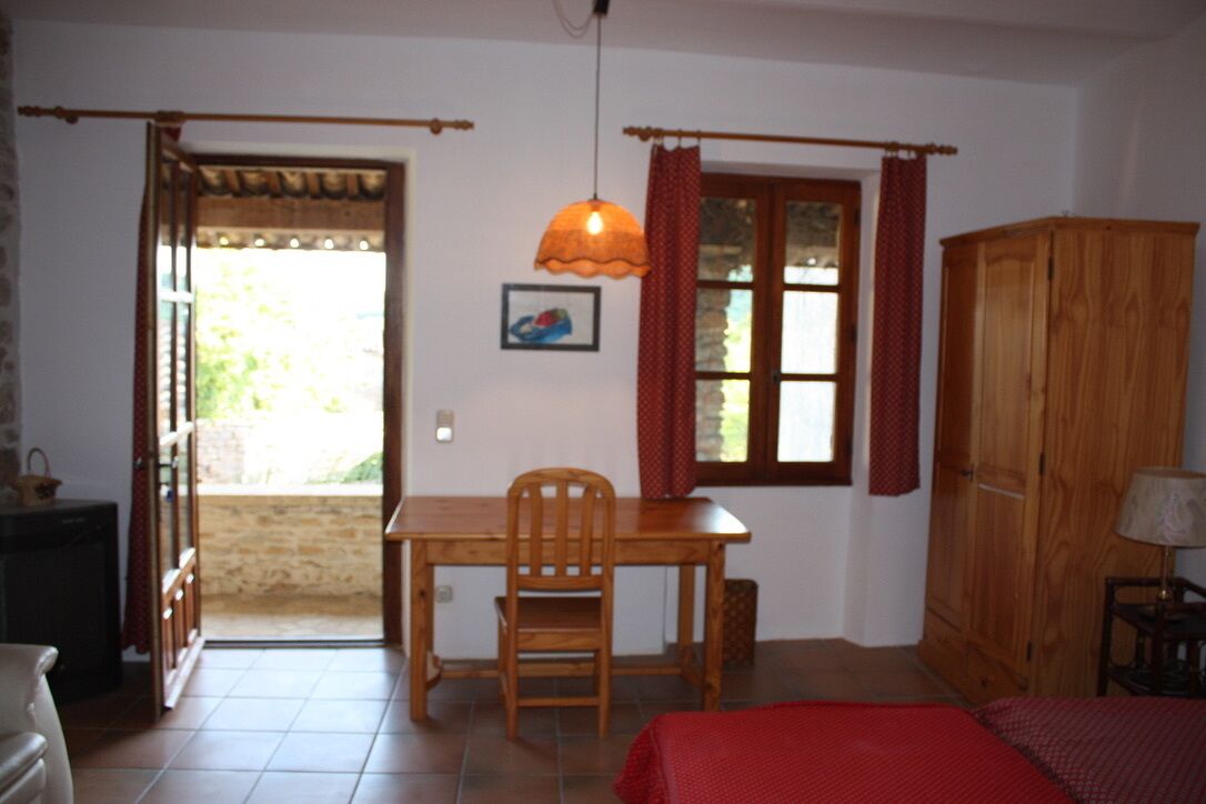 Une pièce avec une table et une chaise en bois, un lit recouvert d’une couverture rouge, une armoire en bois et une fenêtre avec des rideaux.